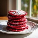 A tall stack of vibrant Red Velvet Pancakes dusted generously with white powdered sugar on a white plate.