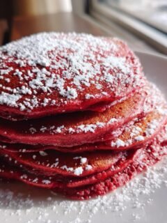 A tall stack of vibrant Red Velvet Pancakes generously dusted with white powdered sugar on a white plate.