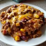 A close-up serving of Slow Cooker Cowboy Casserole featuring ground meat, kidney beans, corn, and melted cheddar cheese on a white plate.