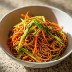 Close-up of a bowl filled with brown Soba Noodle Salad topped with bright julienned carrots and cucumbers, sprinkled with sesame seeds.