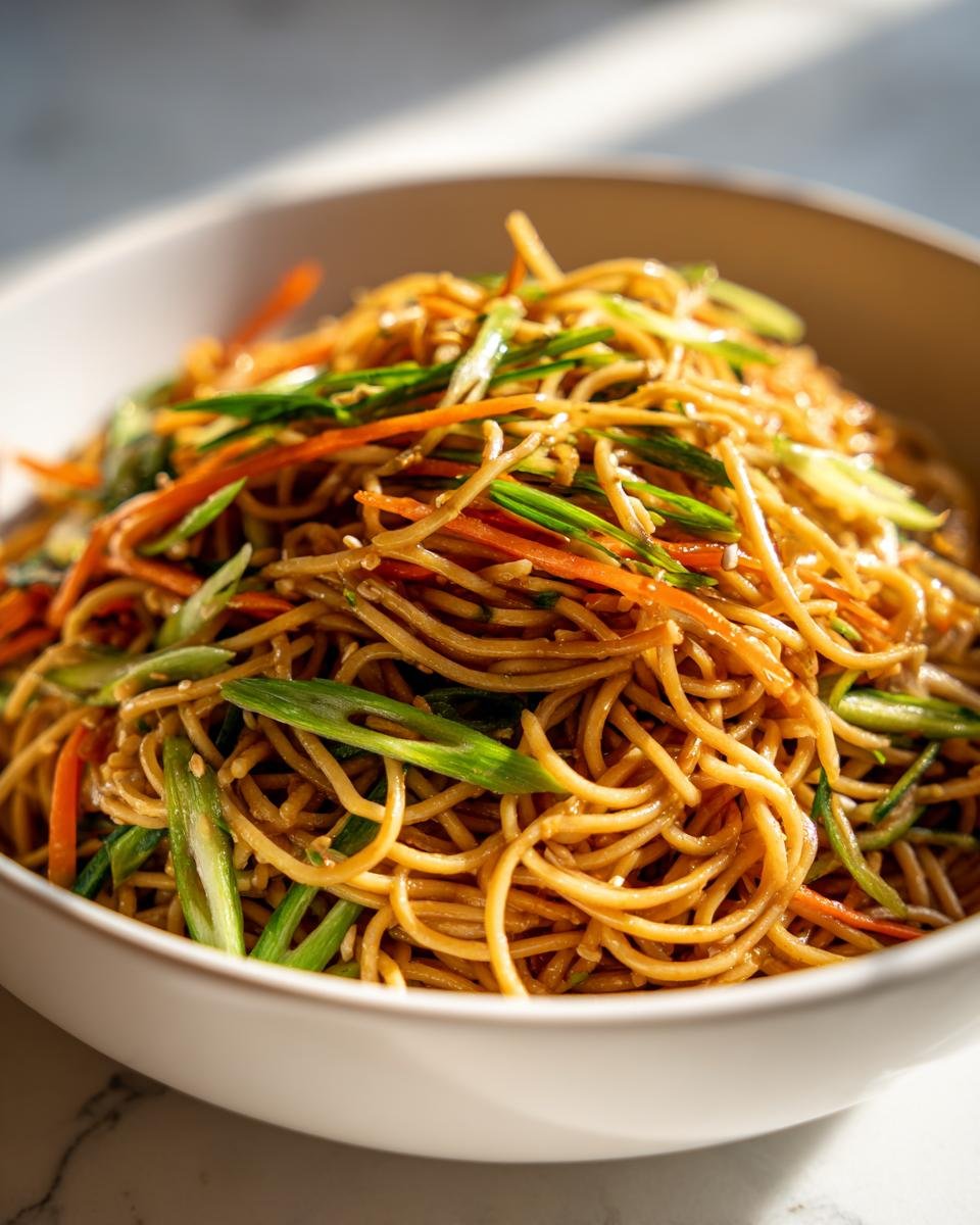 Close-up of a white bowl filled with Amazing Soba Noodle Salad tossed in a savory sauce with julienned carrots and green onions.