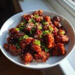 Close-up of crispy Spicy Gochujang Chicken pieces coated in a glossy red sauce, topped with sesame seeds and green onions.