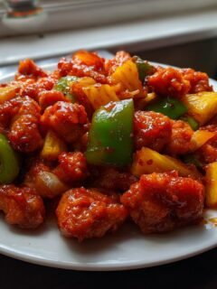 A plate piled high with glistening Sweet And Sour Chicken With Pineapple, featuring crispy chicken pieces, bright pineapple chunks, and green bell peppers.