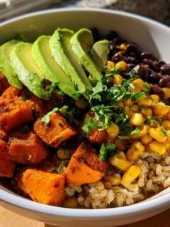 A close-up of a Sweet Potato Burrito Bowl featuring seasoned sweet potato cubes, brown rice, black beans, corn, and sliced avocado.